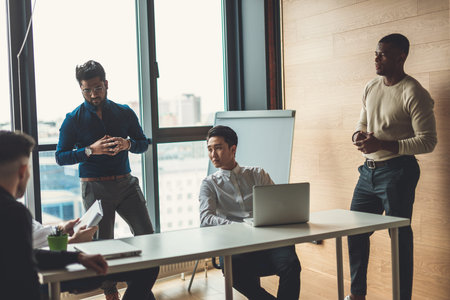 Multiracial male staff of architectural and design bureau communicating in modern loft office with...