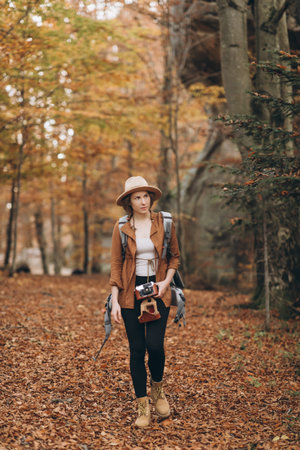 Attractive young woman in a stylish hat and travel bag on her shoulders, looking around at the charming autumn forestの写真素材