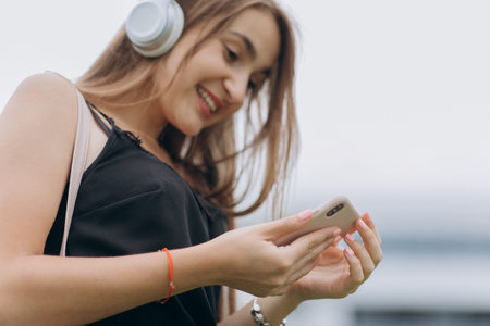 Beautiful young woman using smart phone, typing messages, listening to music, walking on the urban...
