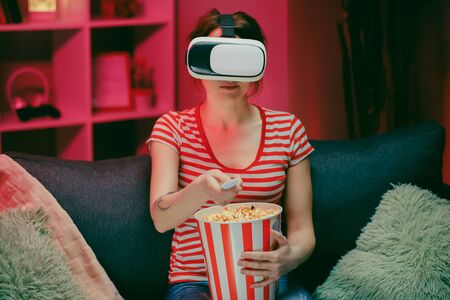 Portrait of the young woman sitting on the couch and having the VR headset, watching something while eating popcorn and smilingの写真素材