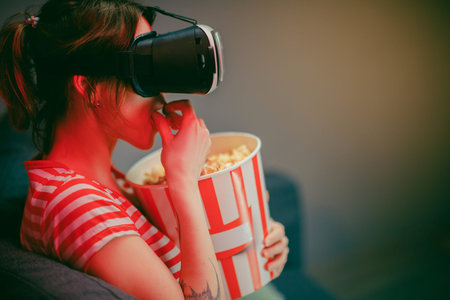Woman wear VR headset and watch movie with popcorn at night. American woman sitting on the sofa in...