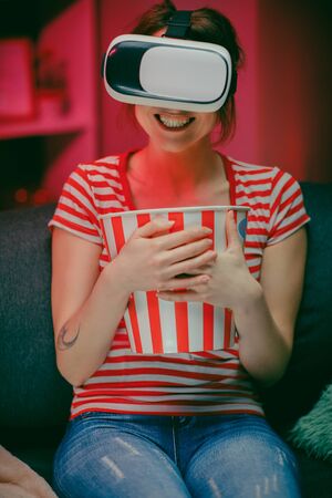Woman wear VR headset and watch movie with popcorn at night. American woman sitting on the sofa in the VR glasses and watching something while eating popcorn.の写真素材