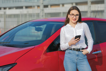 Girl stands with phone near her red electric car and waits when vehicle will charged. Plugging in...