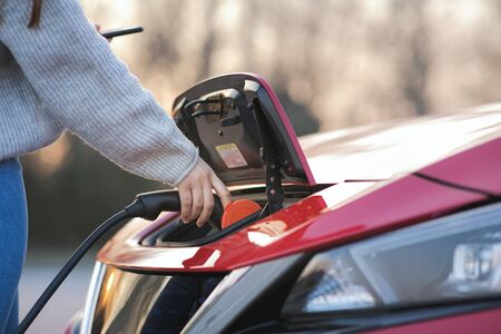 Woman is plugging electric vehicle for charging car battery at parking. Close up. Plugged charging cable electric vehicle, EV parking, power charger cable, charging port station, sustainable futureの写真素材