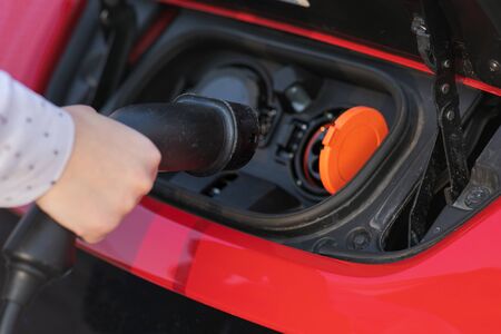 Woman Plugs in Charger into Socket of Her Modern New Red Electric Car. Woman is plugging electric vehicle for charging car battery at parking.の写真素材