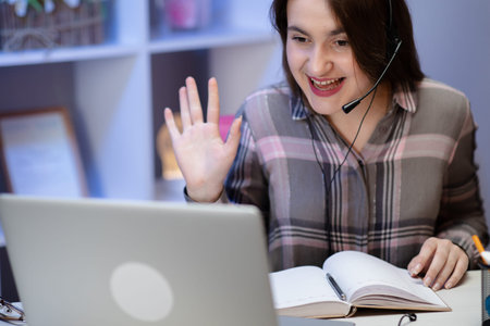Happy woman wearing headphones greeting during a video conference on line with a red laptop sitting...