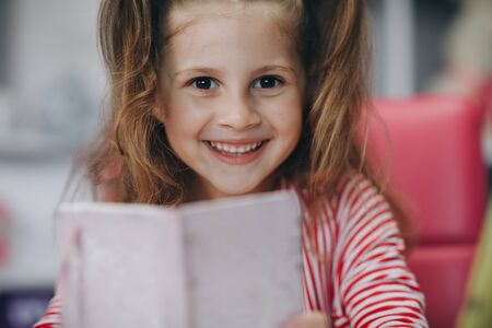 Portrait of a preschool girl looking at the camera and smiling. Kid girl sitting at the desk in children room at home and studying online. Distance learning, school onlineの写真素材