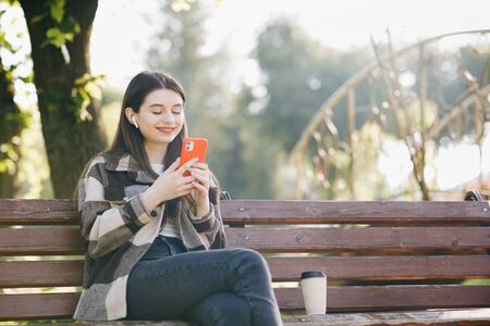 Portrait of girl wearing earphones and using smartphone listening music. Attractive woman browsing on mobile phone in park. City, urban backgroundの写真素材