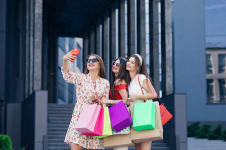 Group of young beautiful girls in casual clothes with sunglasses, makeup, hair hoop and colored shopping bags making selfie after successful shopping. Building And Park Plants At The Backgroundの写真素材
