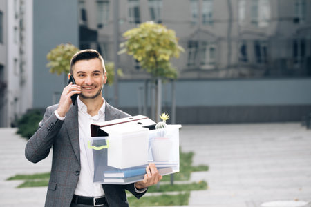 Cellphone conversation. An employee between office buildings with a box and documents with a desk flower. Happy fired man talking on phone with stuff in box. Speaking on telephoneの写真素材
