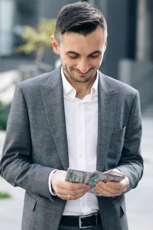 Handsome Rich Man Wearing Stylish Suit Counting Money Standing in the Street Near Office Buildingの写真素材