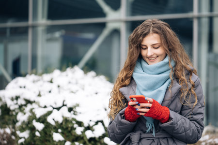 Curly-haired female texting on smartphone standing on street in winter city. Female tapping on cellphone outdoors. Vacation winter outdoor. Happy Young Woman Enjoys Life.の写真素材