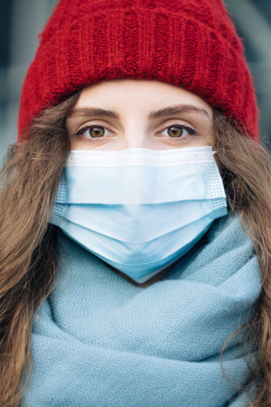 Close up portrait of Caucasian young curly female in medical mask standing in winter city. Crowd people sick covid-19. Epidemic coronavirus. Pandemic flu corona virusの写真素材