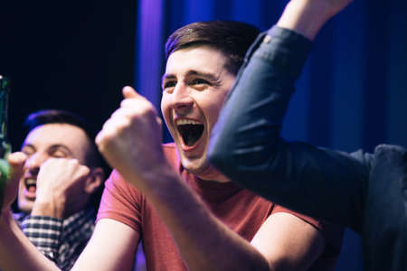 Close up of Caucasian male football fan sitting with friends at TV screen in dark living room and watching match. Cheerful man celebrating football win. Scoring goal or winning game conceptの写真素材