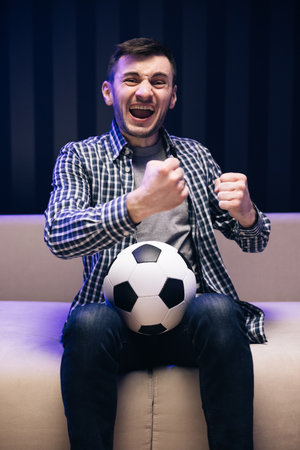 Gladly Surprised Reaction Of A Young Man Is Watching Tv And Sitting On The Sofa. Sports Fan Reaction Concept. Excited sports fan watching his favourite player on TV. Football fan watching championshipの写真素材
