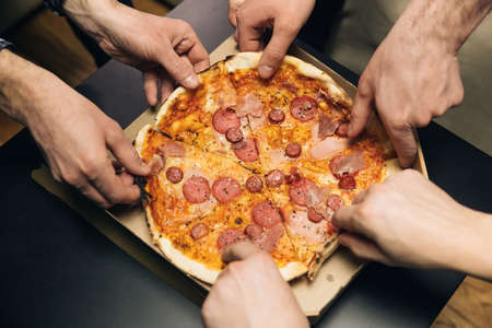 Male hands taking slices of pizza with cheese, tomatoes and ham from food delivery. Group of hungry friends sitting at desk and sharing delicious lunch on table backgroundの写真素材
