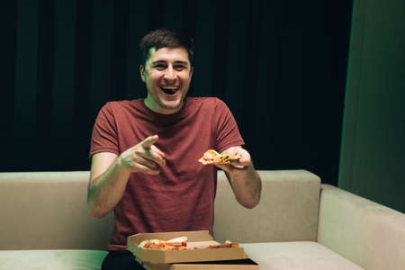 Portrait of young hipster eating pizza, watching favorite comedy on tv while relaxing at sofa in the living room. Happy man eating Italian food.の写真素材
