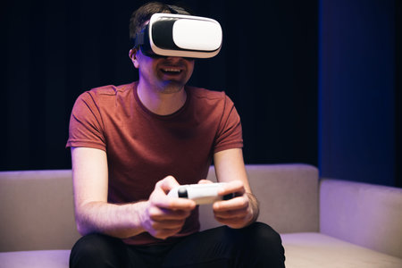 Attractive caucasian man hold joystick and wear virtual reality glasses headset helmet on sofa at...