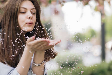 Woman Enjoying Spring Blossom. Happy Smiling Girl blowing sakura flower petals, making them fly to camera. Sunny Spring Outdoors Activity. Spring blossom of sakura tree in city park.の写真素材