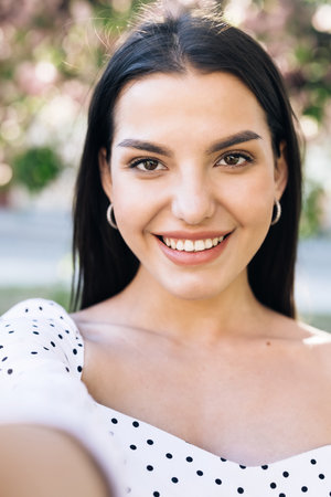 Young smiling woman in summer white dress taking selfie self portrait photos on smartphone. Model posing on park sakura trees background. Female showing positive face emotions.の写真素材