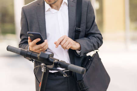 Unrecognizable businessman in suit is coming up to an electric scooter. Phone application for rental scooter. Ecological city transport. Businessman rents electric scooter using mobile phone appの写真素材
