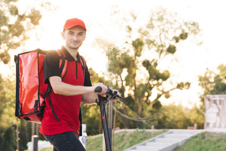 Young student works in a delivery service, with a red backpack and baseball cap, rides an electric scooter along the embankment at sunsetの写真素材