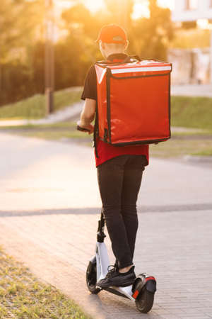 Unrecognizable young courier rides on his e-scooter. Deliver male parcel delivery service guy rides an electric scooter down an empty sidewalk with red backpack down the street at sunsetの写真素材