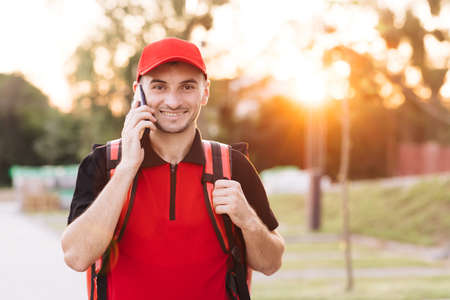 Food delivery guy with red backpack deliver orders. Male courier with isothermal food case box on electric scooter arrives to the entrance to the house and calls for clientの写真素材