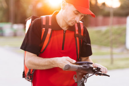 Man courier food delivery with red thermal backpack walks street with electric scooter uses...