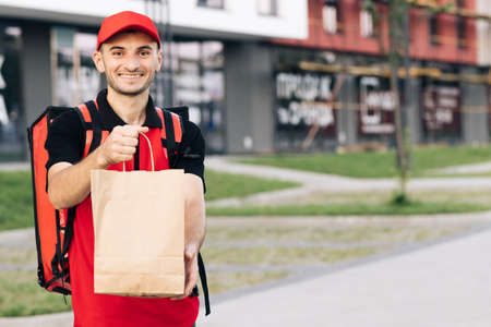 Happy young courier is proud of his job smiling standing in the street. Home delivery. Outdoor portrait of delivery man with red uniform holding food bags waiting for customerの写真素材