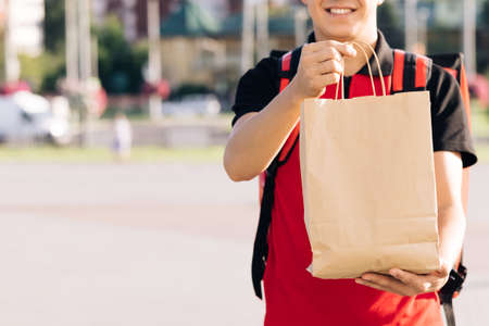 Close up portrait of positive young man courier person. Delivery service door to door. Happy delivery worker holding packet with food looking at the camera and smilingの写真素材