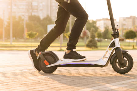 Shot on legs young man in black sneakers ride on electric mobile scooter. Close up feet man courier food delivery rides the street on electric scooter deliver online order client at sunsetの写真素材