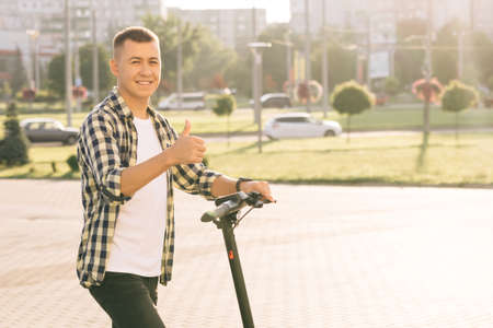 Portrait shot of caucasian hipster man leaning on electric scooter looking at camera and showing thumb up. Hipster man on vehicle outdoors. Eco-friendly modern urban transportの写真素材
