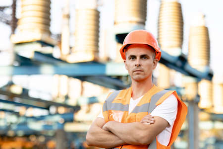 Portrait of engineer worker in uniform and helmet standing near high voltage substation with tall pylons and voltage distribution cablesの写真素材