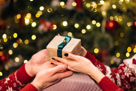 Close-up hands of man giving festive box with Christmas present to woman sitting at dinner feast table during holiday family party, decorating xmas tree and celebration lights, selective focusの写真素材