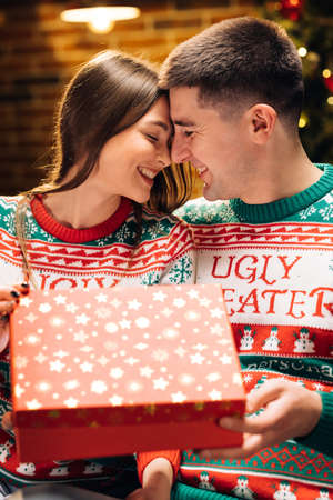 Happy man is making christmas gift to his beloved woman. The woman is surprised and excited after opening received gift box. Concept of holidays, romance, surprise, e-commerce, Xmas, Holiday miracleの写真素材