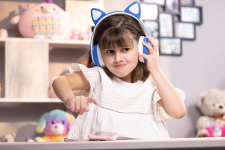 Small kid girl in headphones using funny editing application on smartphone, enjoying cool video or music content in social network, playing online games, communicating distantlyの写真素材