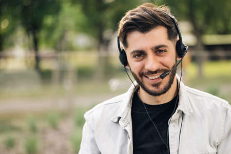 Close up of Face young handsome man operator look at camera smile taking calls smile outdoor call center office business worker computer corporate headset job service group finance phone.の写真素材