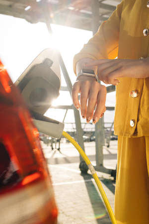 Close up of woman hands attaching power cable supply to charge electric or EV car using app on wearable smart watch. Female plugging an electric car or EV at electric charging stationの写真素材