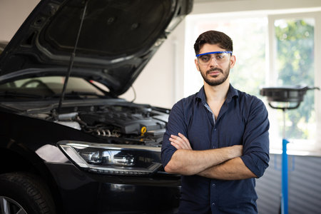 Portrait Bearded Handsome Car Mechanic is Posing in a Car Service. He Wears a Jeans Shirt and Safety Glasses. His Arms are Crossed. Specialist Manager Looks at a Camera and Smilesの写真素材