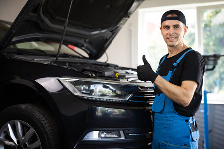 Positive auto service worker smiling to camera and showing thumb up gesture, approving car repair workshop. Portrait of car mechanic in a car workshop shows thumbs up, in the background of serviceの写真素材