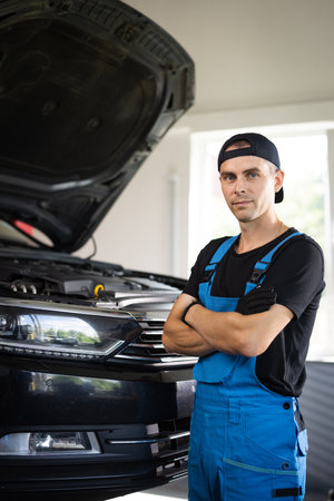 Portrait shot of caucasian man in blue uniform and hat standing in big auto garage, looking to camera and crossing hands. Pretty male mechanic in maintaining serviceの写真素材