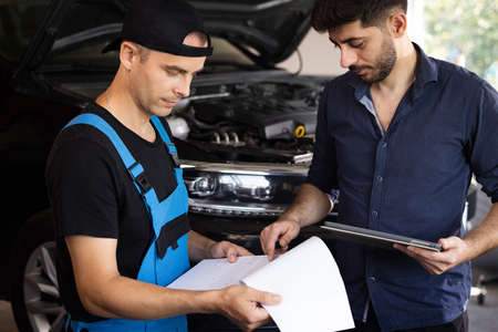 Car Service Employees Inspect the Bottom and Skid Plates of the Car. Manager Checks Data on a Tablet Computer and Explains the Breakdown to Mechanic. Specialist is Showing Info on Tablet.の写真素材