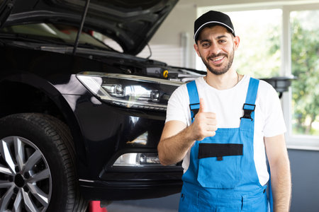 Portrait of bearded car mechanic in a car workshop shows thumbs up. Positive auto service worker in blue overalls and cap smiling to camera and showing thumb up gesture, approving car repair workshopの写真素材