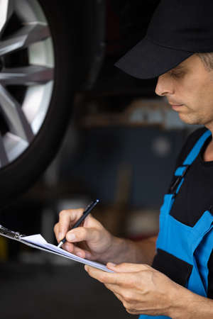 Car service employe inspect car. Mechanic inspects the car undercarriage way and makes a note on his inspection sheet. Automobile service, car mechanic. Modern workshop.の写真素材