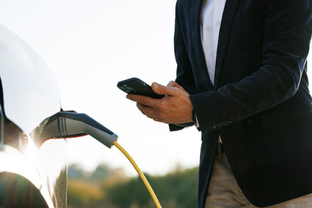 Unrecognizable man plugging electric car from charging station. Male plugging in power cord to electric car using app on smartphone. Businessman charging electric car at outdoor charging statioの写真素材