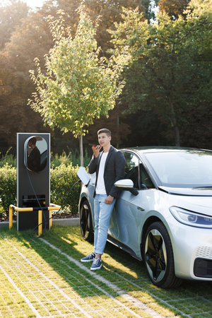 Caucasian man using smartphone voice messages and waiting power supply connect to electric vehicles for charging the battery in car. Plug charging an electric car from charging stationの写真素材