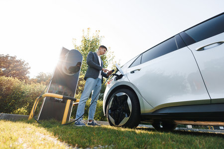 Man plugging electric car from charging station. Male attaching in power cord to electric car using app on smartphone. Businessman charging electric car on charging station at sunset.の写真素材