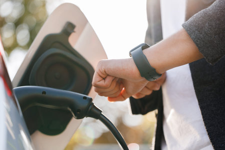 Unrecognizable man disconnects charging cable to to electric vehicle using smartwatch. Male hand...