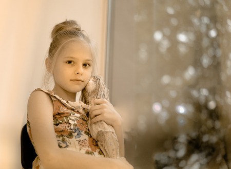 vintage, children. retro style. Pretty blond girl with a doll on the background of a large window with lightsの写真素材